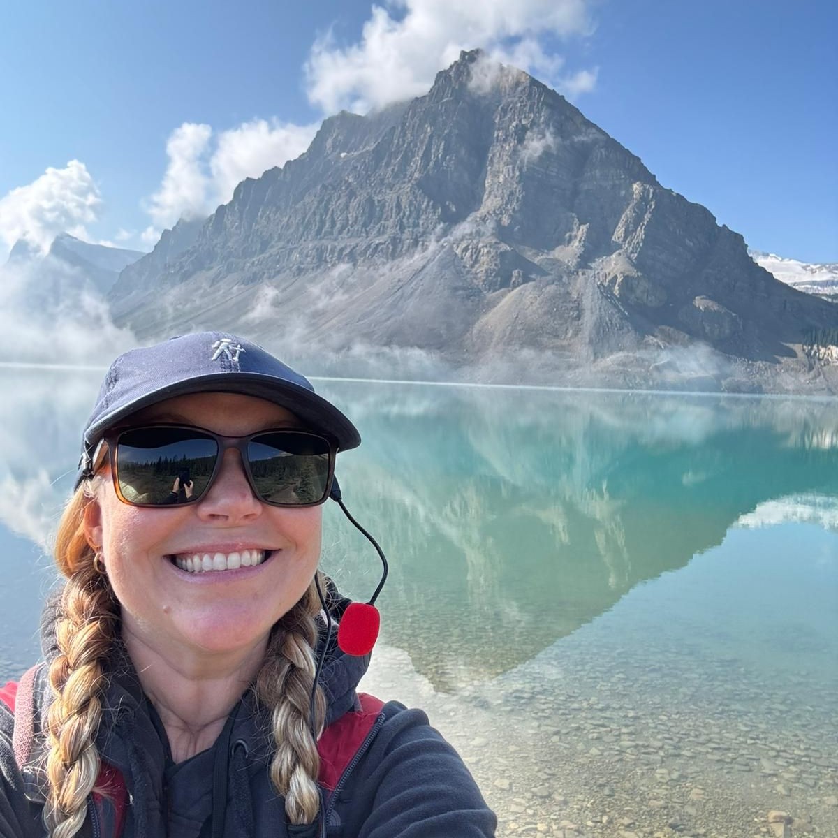 Smiling woman in cap and sunglasses takes a selfie with a jagged mountain and turquoise lake in the background.