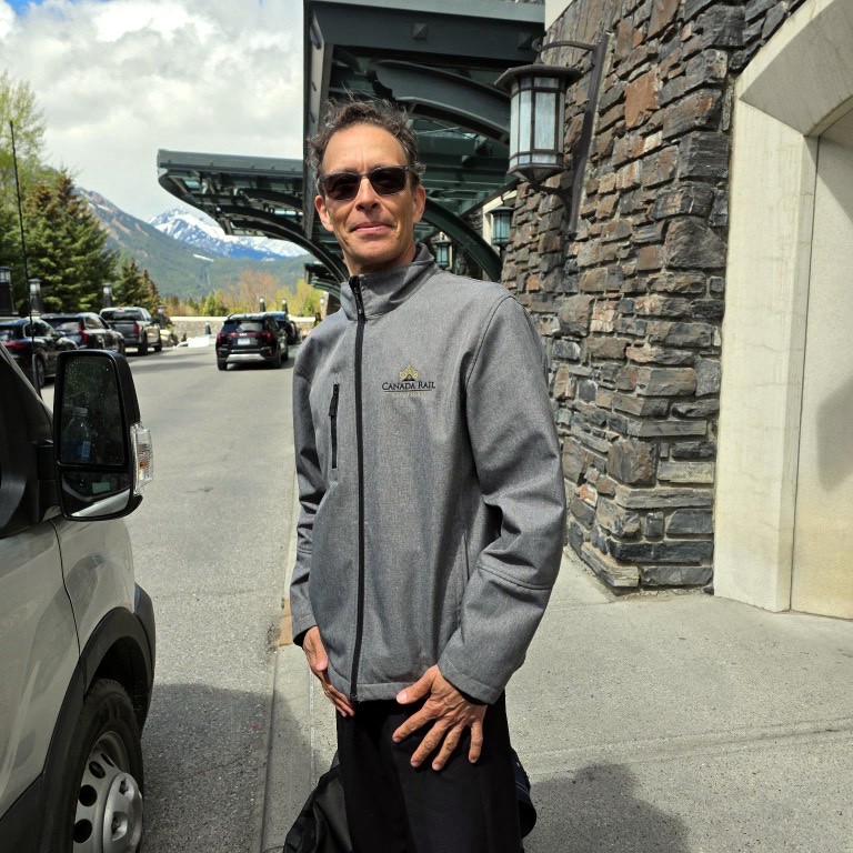 Man in a gray Canada Rail jacket stands outside a stone building with mountains in the background, wearing sunglasses.